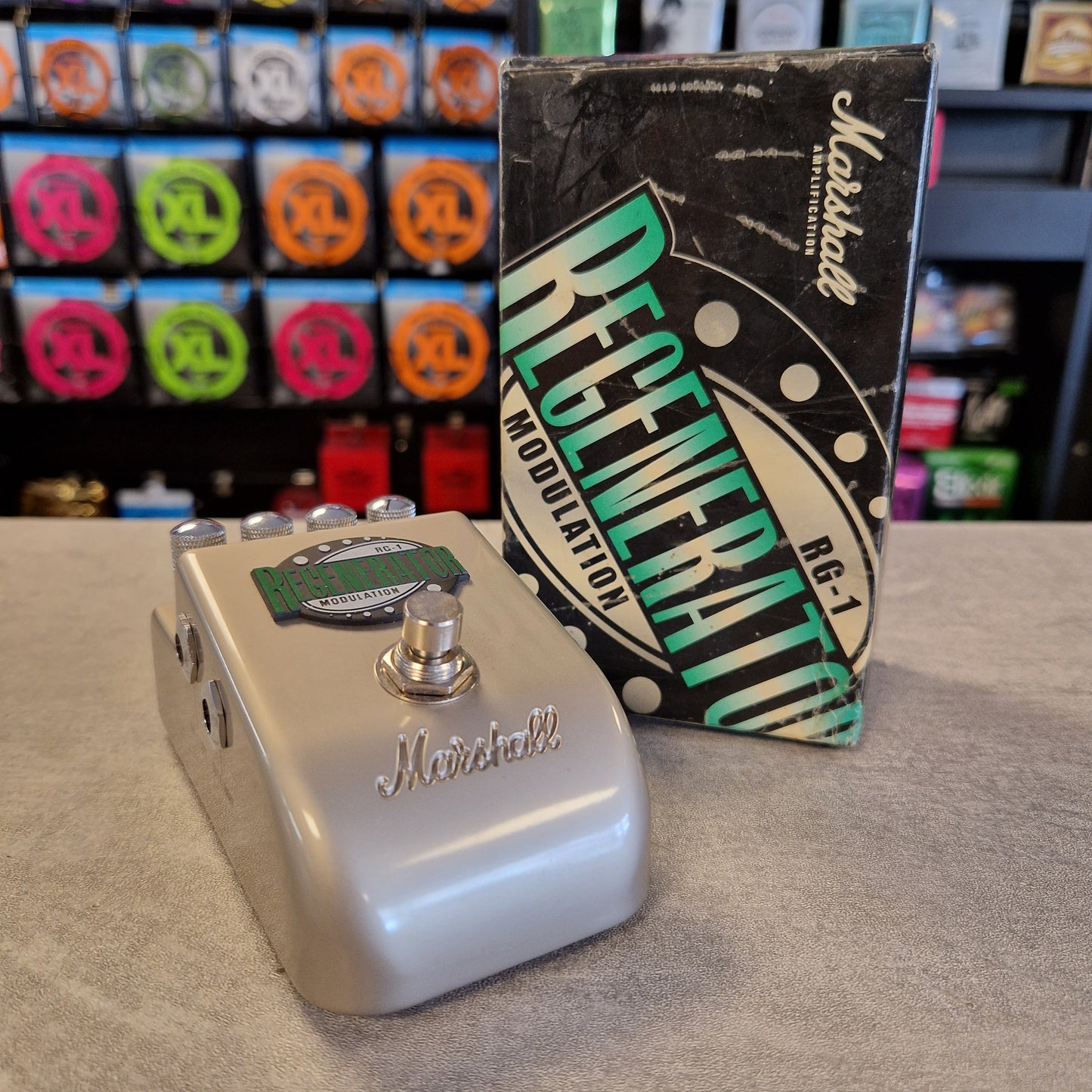 Pre-Owned Marshall RG1 Regenerator Modulation Pedal A Marshall Regenerator RG-J modulation guitar pedal in a metallic silver finish, shown with its original black and green packaging box on a gray tabletop. The background features a blurred shop display of Marshall speakers.