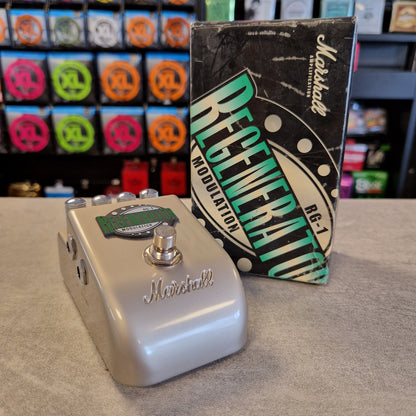 Pre-Owned Marshall RG1 Regenerator Modulation Pedal A Marshall Regenerator RG-J modulation guitar pedal in a metallic silver finish, shown with its original black and green packaging box on a gray tabletop. The background features a blurred shop display of Marshall speakers.