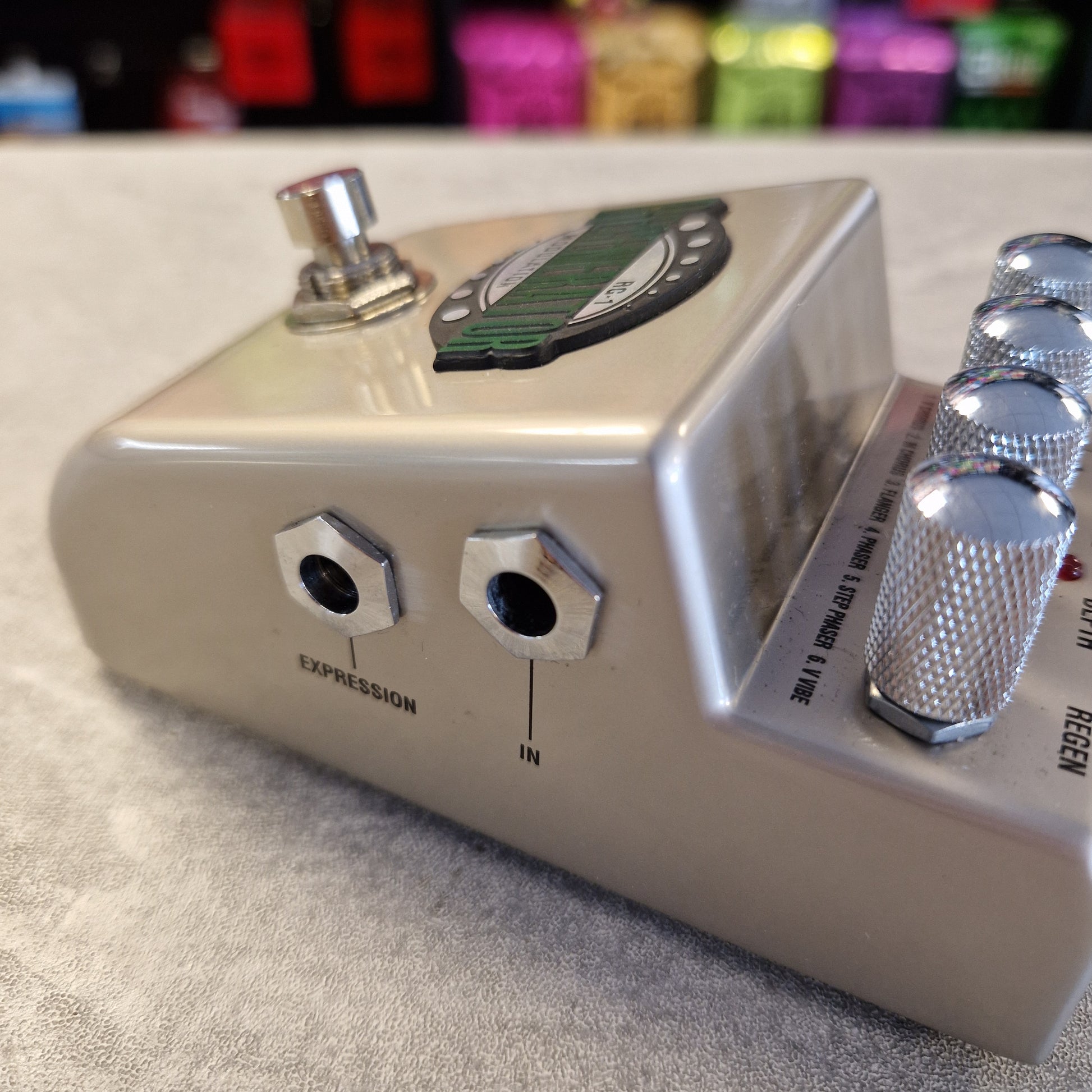 Pre-Owned Marshall RG1 Regenerator Modulation Pedal A close-up shot of a silver metal guitar effects pedal, showing its footswitch, expression and input jacks, and control knobs. The pedal is resting on a textured gray surface with colorful, blurred background items.