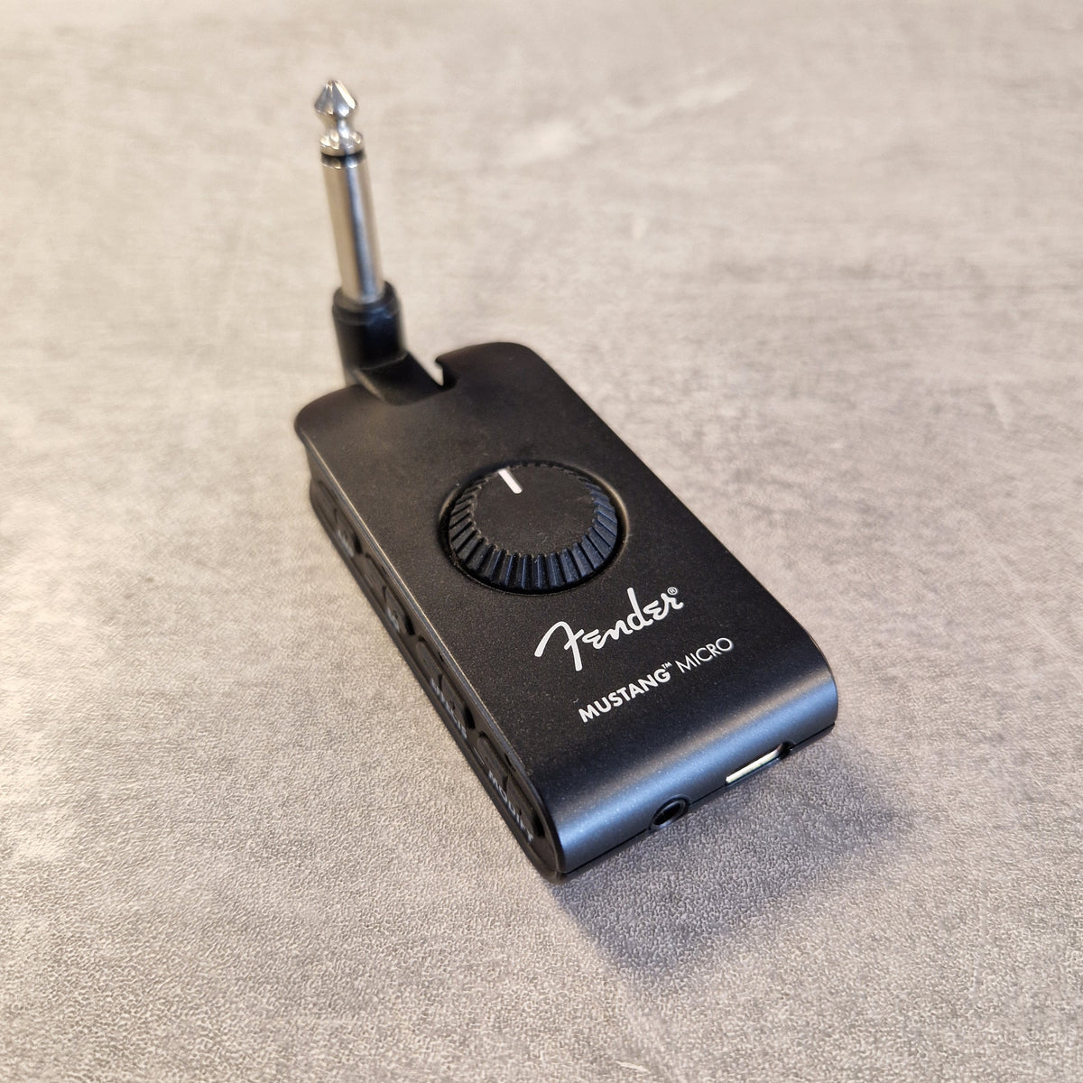 Pre-Owned Fender Mustang Micro Headphone Guitar Amplifier A Fender Mustang Micro wireless guitar amplifier receiver with a built-in 1/4-inch jack and adjustable volume knob, shown on a textured gray surface.