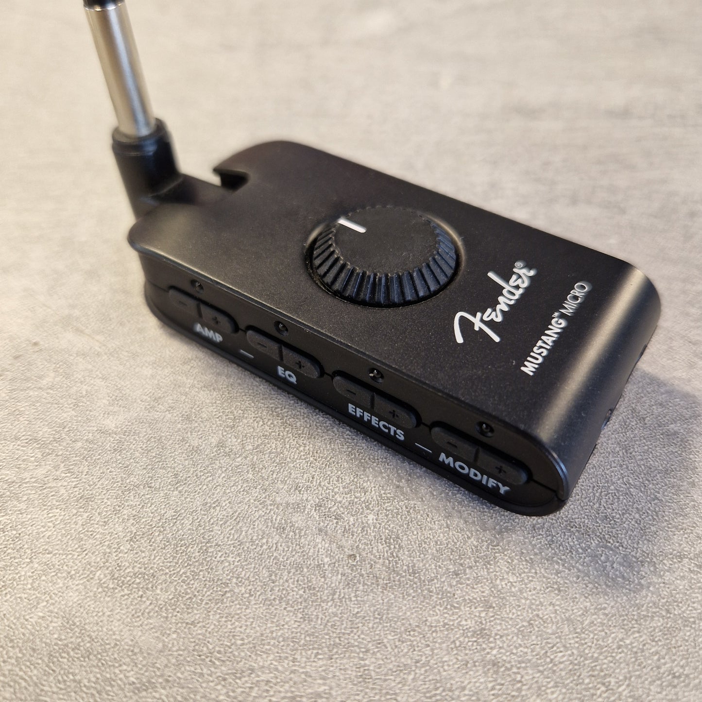 Pre-Owned Fender Mustang Micro Headphone Guitar Amplifier A black Fender Mustang Micro wireless guitar amplifier receiver with a metal antenna, resting on a textured gray surface.