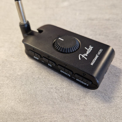 Pre-Owned Fender Mustang Micro Headphone Guitar Amplifier A black Fender Mustang Micro wireless guitar amplifier receiver with a metal antenna, resting on a textured gray surface.