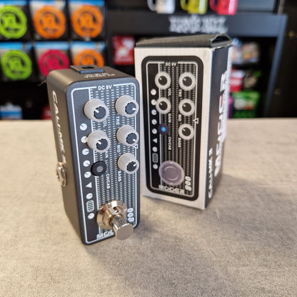 Pre-Owned Mooer 008 Cali-Mk3 Dual Channel Preamp Pedal A black and gray modulation pedal with white text and knobs, standing next to its original retail box on a gray tabletop. The background shows a store shelf with colorful items.