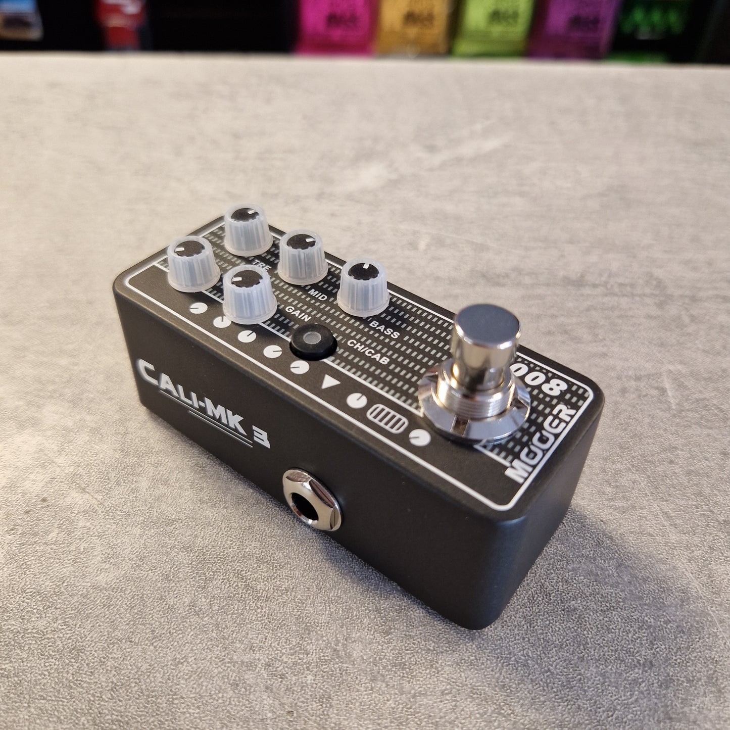 Pre-Owned Mooer 008 Cali-Mk3 Dual Channel Preamp Pedal A Call of Mk III Megaphone guitar effect pedal with a black metal body, white and silver knobs, and a silver footswitch, resting on a gray textured surface.
