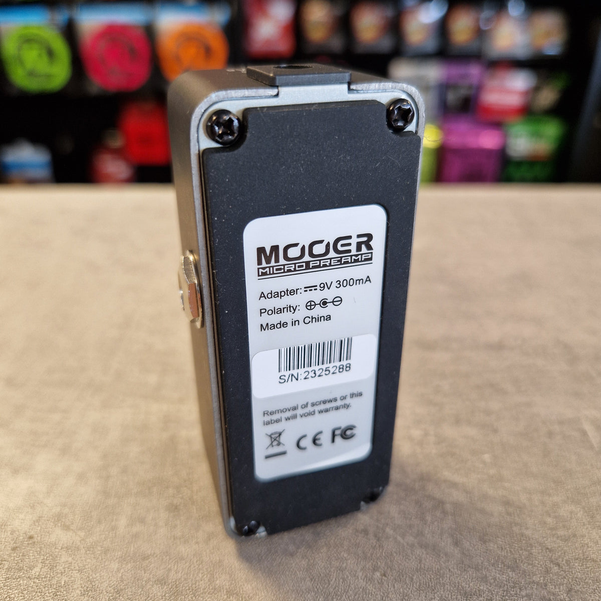 Pre-Owned Mooer 008 Cali-Mk3 Dual Channel Preamp Pedal A MOOGER Mini Amp DC power adapter, 9V 300mA, shown from the back side with its white product label. The adapter has a black plastic body with a silver metal trim and corner screws, resting on a beige surface with a blurred background.