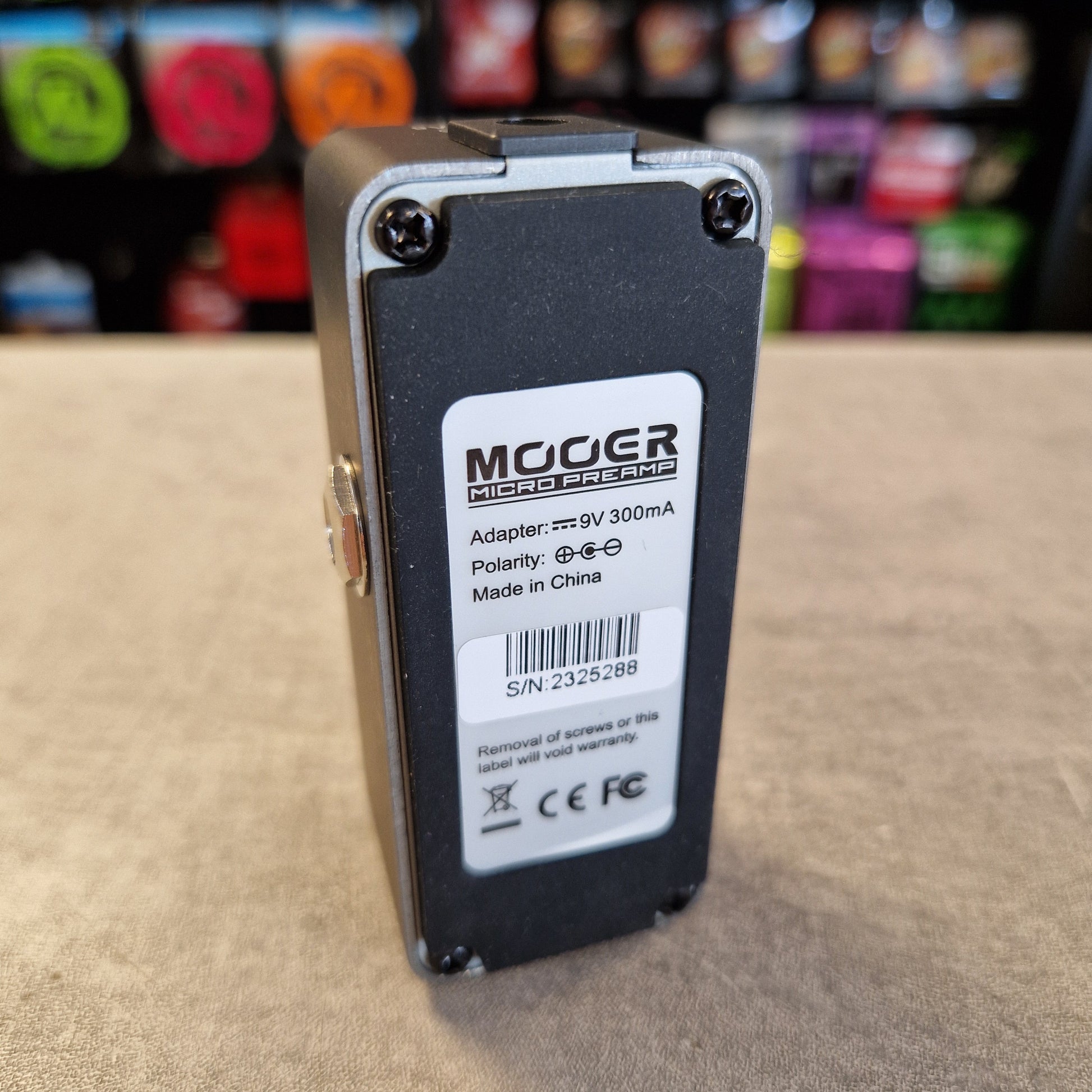 Pre-Owned Mooer 008 Cali-Mk3 Dual Channel Preamp Pedal A MOOGER Mini Amp DC power adapter, 9V 300mA, shown from the back side with its white product label. The adapter has a black plastic body with a silver metal trim and corner screws, resting on a beige surface with a blurred background.