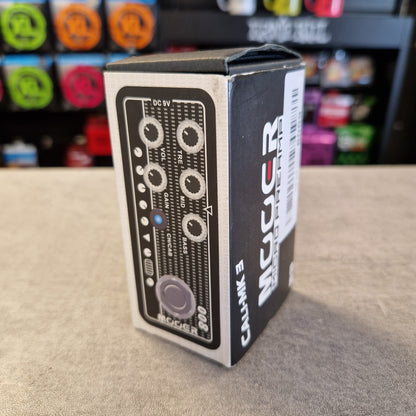 Pre-Owned Mooer 008 Cali-Mk3 Dual Channel Preamp Pedal A boxed audio device labeled 'MUGLER' and 'CAJAK 3' standing upright on a gray tabletop, with a blurred store background behind it.