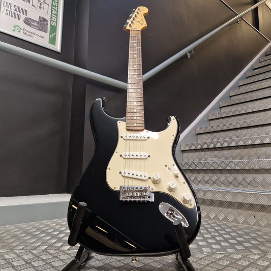Pre-Owned Fender Standard Mexican Stratocaster 2011 Electric - Black A black Stratocaster-style electric guitar with a cream pickguard and maple neck, displayed on a stand in front of an indoor staircase.