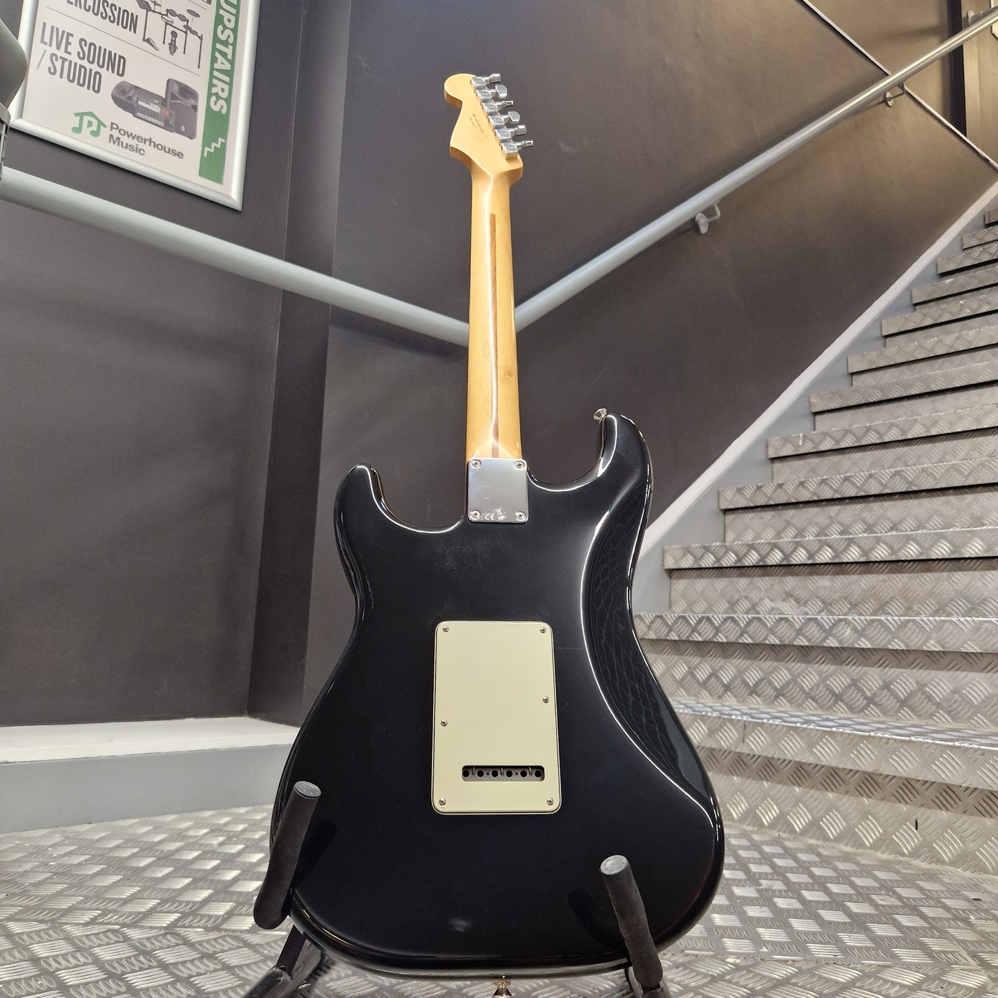 Pre-Owned Fender Standard Mexican Stratocaster 2011 Electric - Black A black electric guitar with a natural wood neck and cream pickguard, displayed on a stand in front of a staircase.