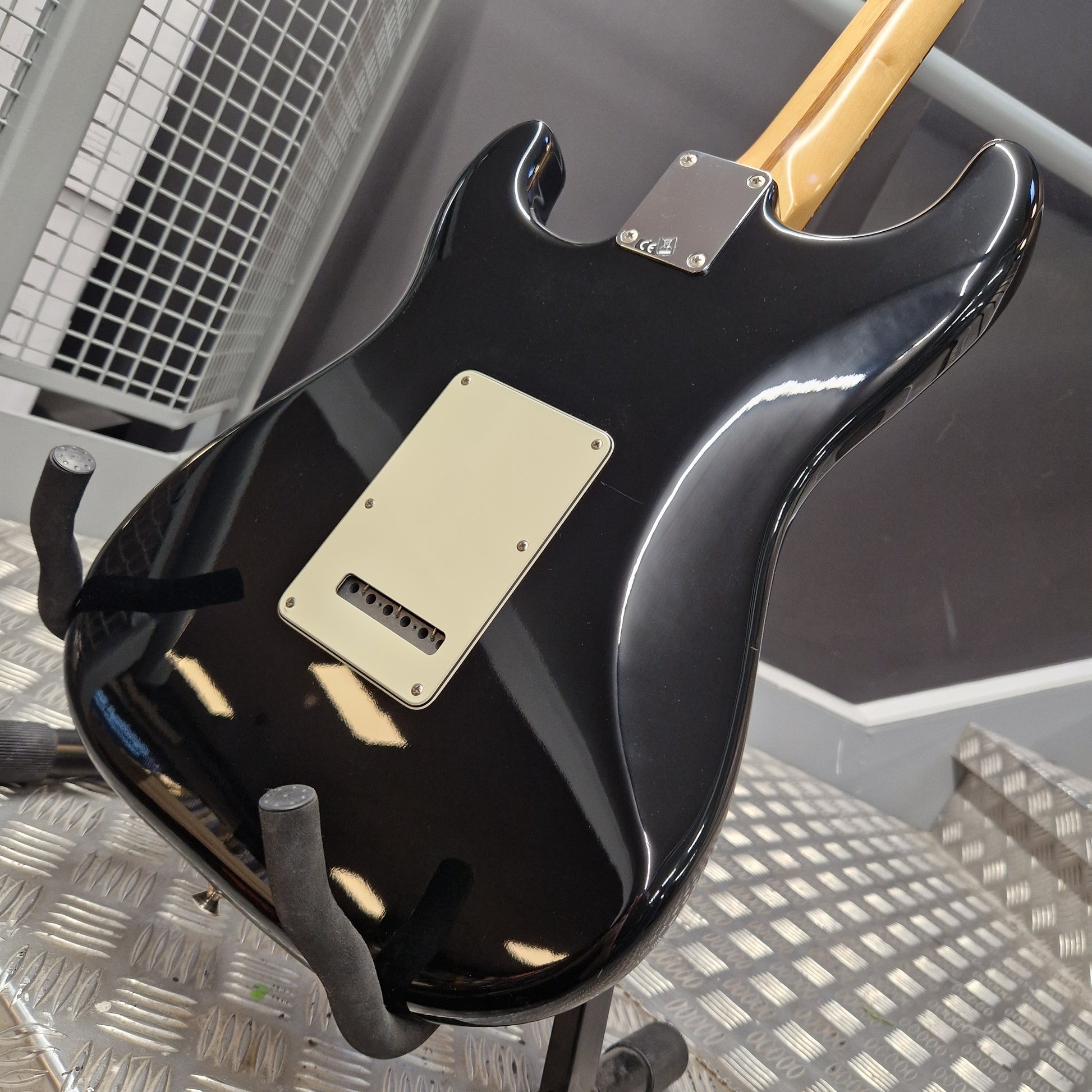 Pre-Owned Fender Standard Mexican Stratocaster 2011 Electric - Black A glossy black electric guitar body, likely a Stratocaster-style model, shown from the back on a stand. It features a cream-colored pickguard and a light natural wood neck visible at the top, photographed in a workshop or garage setting with a metal grate floor.