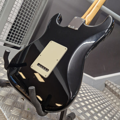 Pre-Owned Fender Standard Mexican Stratocaster 2011 Electric - Black A glossy black electric guitar body, likely a Stratocaster-style model, shown from the back on a stand. It features a cream-colored pickguard and a light natural wood neck visible at the top, photographed in a workshop or garage setting with a metal grate floor.