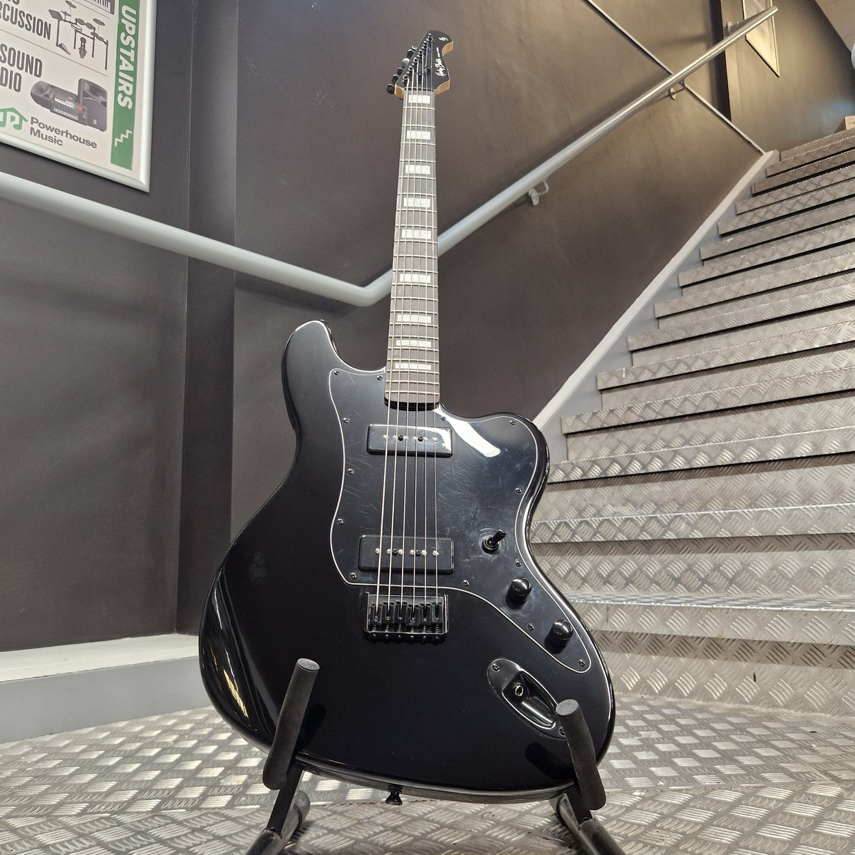 Pre-Owned Harley Benton JA-Baritone HH Electric - Black A black electric guitar with a glossy finish stands on a stand in an indoor stairwell area. The guitar has a double-cutaway body shape, a dark fretboard, and chrome hardware.