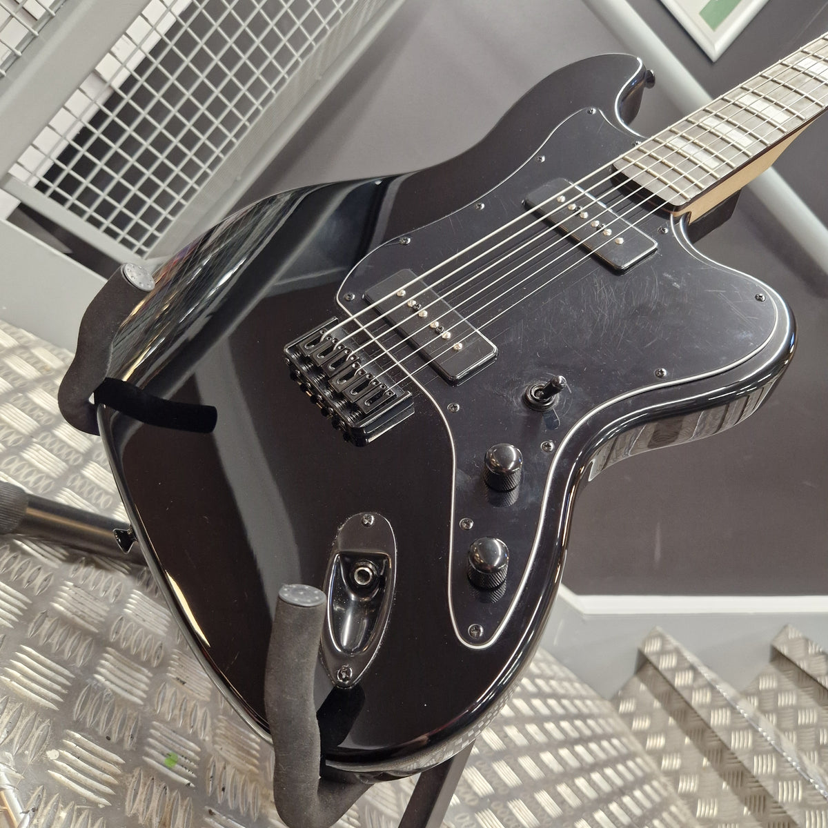 Pre-Owned Harley Benton JA-Baritone HH Electric - Black A glossy black electric guitar with chrome hardware, photographed from an angle on a metal stand in what appears to be a showroom or workshop.
