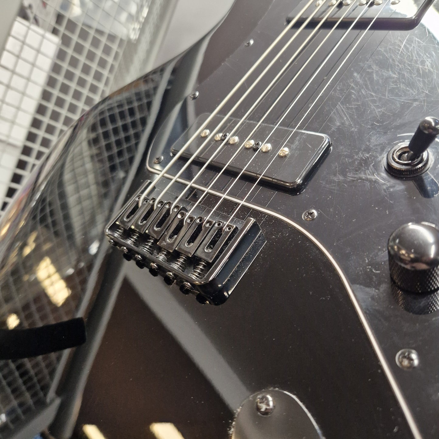Pre-Owned Harley Benton JA-Baritone HH Electric - Black Close-up of a black electric guitar body showing the bridge, pickups, control knobs, and part of the neck, photographed indoors with a blurred grid-patterned background.
