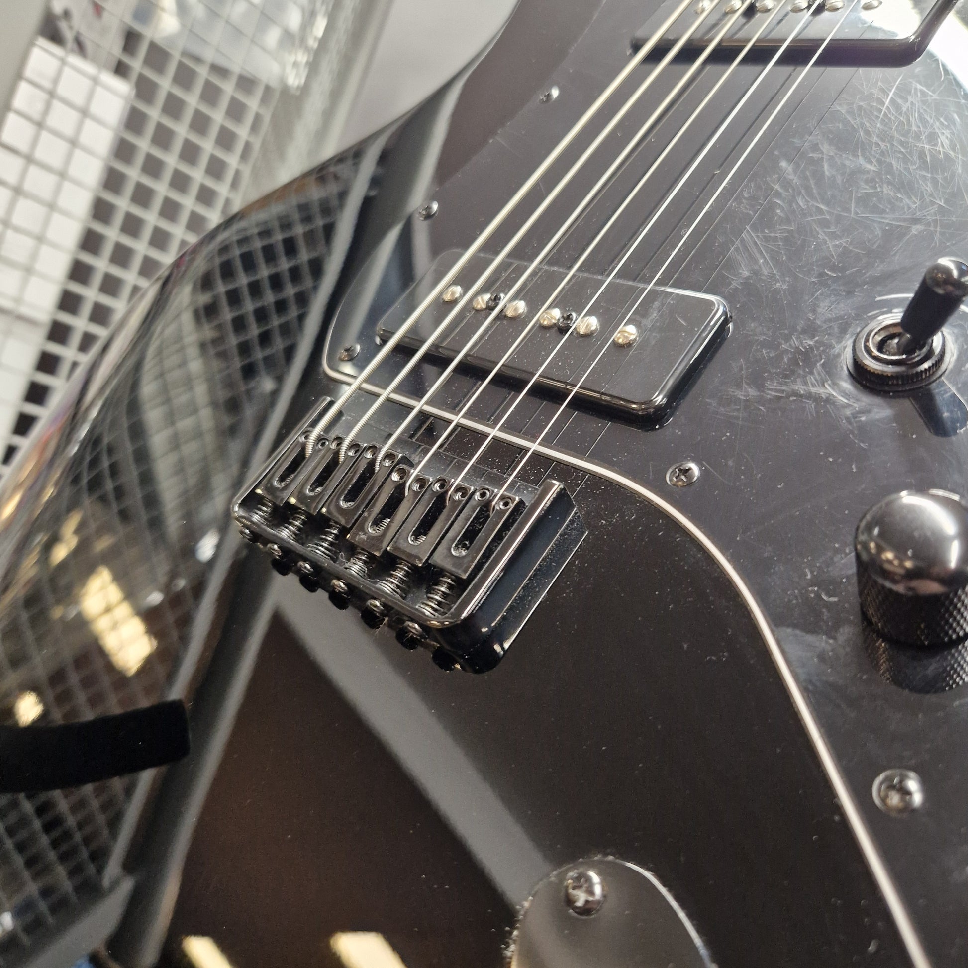 Pre-Owned Harley Benton JA-Baritone HH Electric - Black Close-up of a black electric guitar body showing the bridge, pickups, control knobs, and part of the neck, photographed indoors with a blurred grid-patterned background.