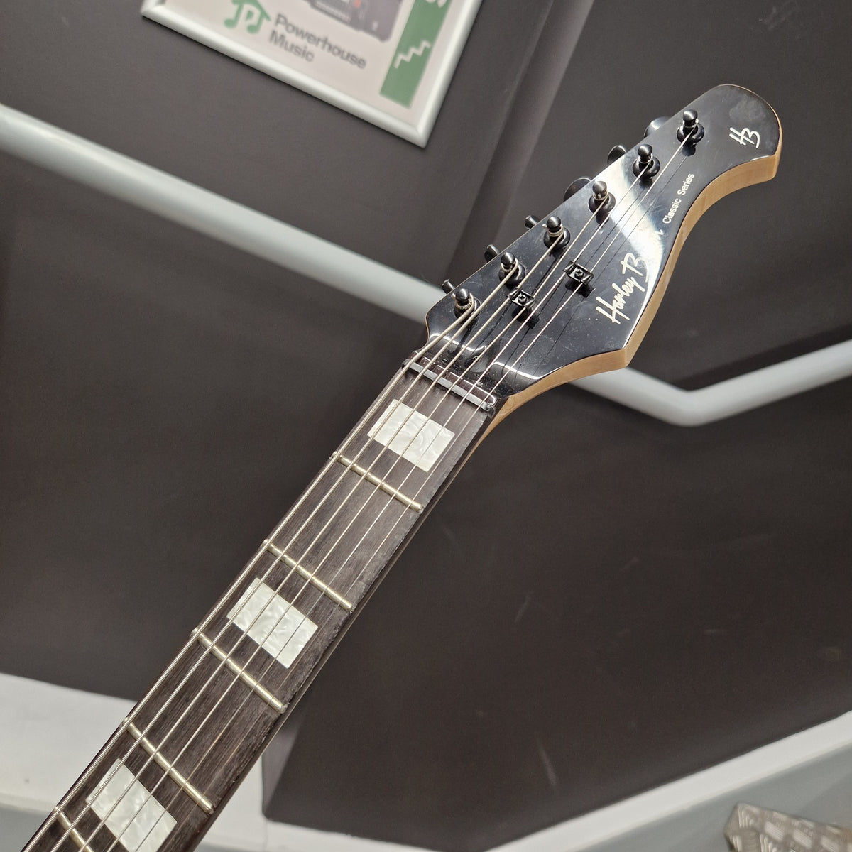 Pre-Owned Harley Benton JA-Baritone HH Electric - Black Close-up of a guitar headstock and neck, showing the Helly Hansen logo and six tuning pegs against a dark gray background.
