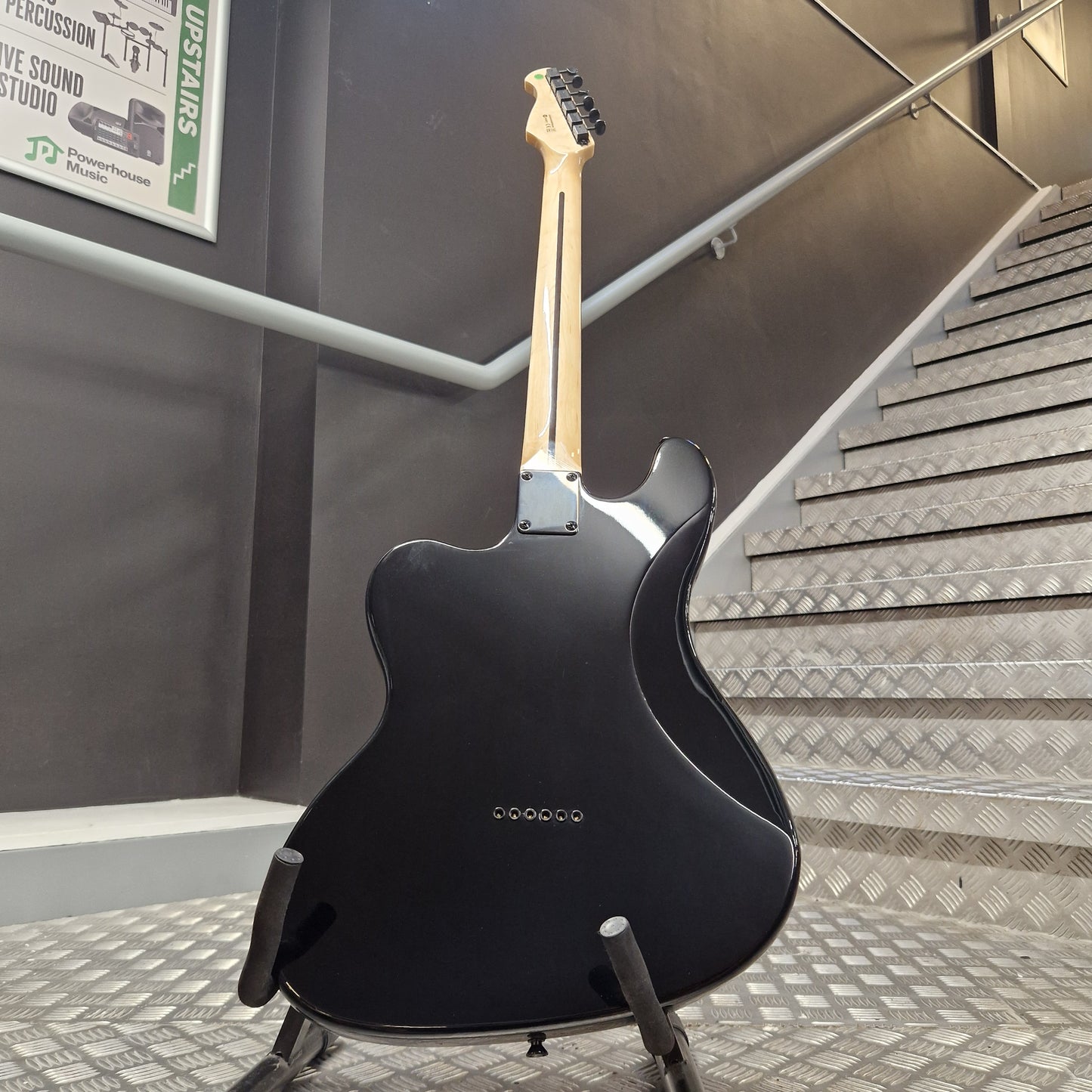 Pre-Owned Harley Benton JA-Baritone HH Electric - Black A black electric guitar body on a stand in an indoor stairwell, with a metal handrail and wall visible in the background.