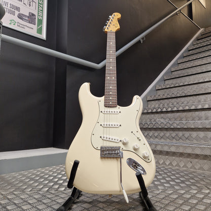 Pre-Owned Fender Standard Strocaster 2014 Electric - Arctic White An off-white electric guitar with a dark brown neck and black pickguard, displayed on a stand in front of a staircase.