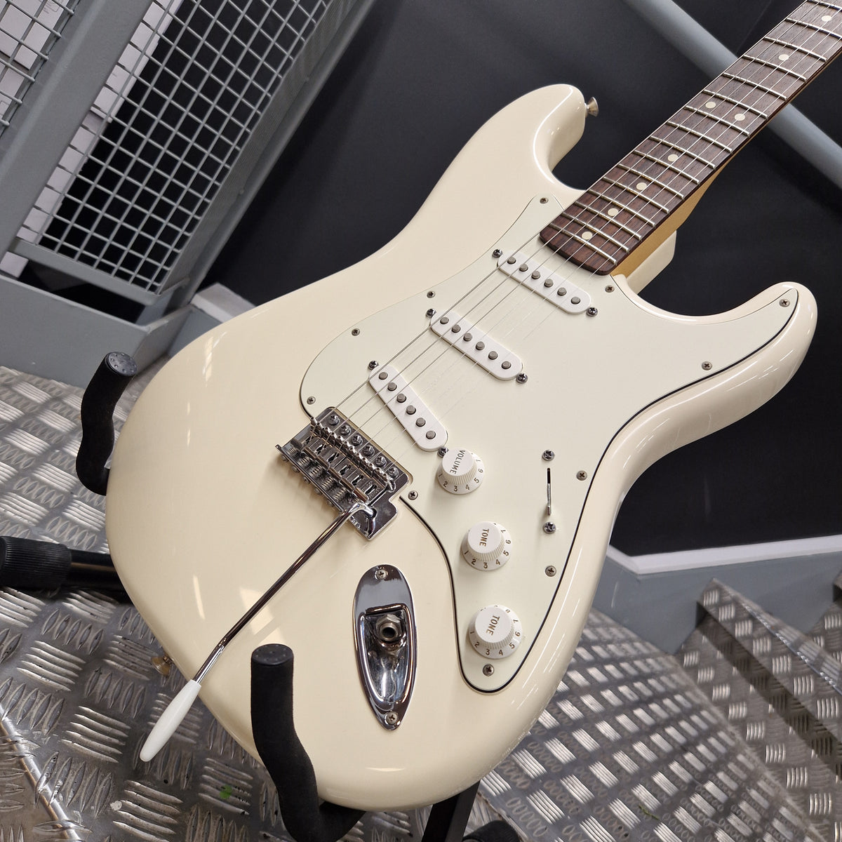 Pre-Owned Fender Standard Strocaster 2014 Electric - Arctic White Cream-colored electric guitar with chrome hardware, resting on a black stand on a textured metal floor.
