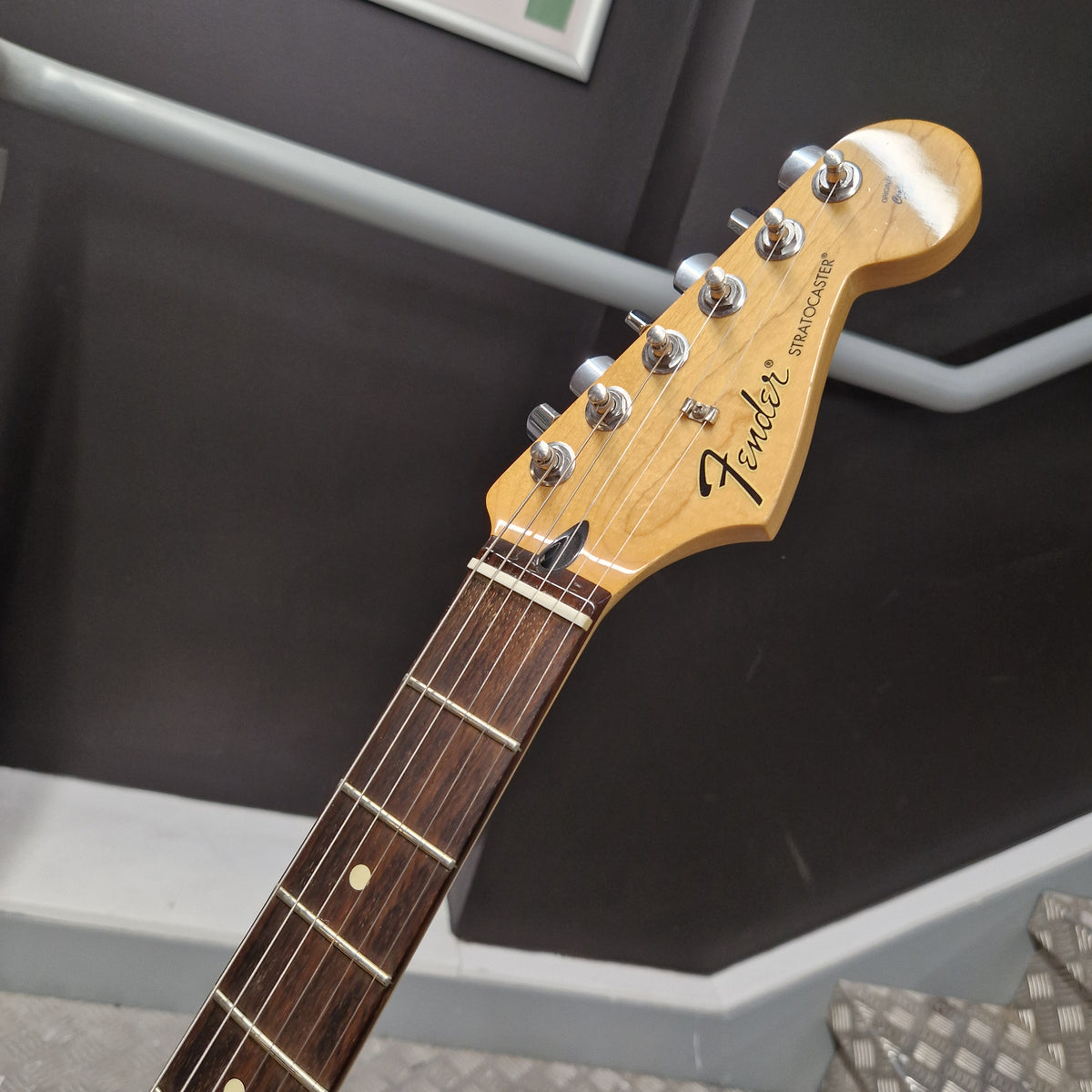 Pre-Owned Fender Standard Strocaster 2014 Electric - Arctic White Close-up of a Fender Stratocaster electric guitar headstock and upper neck, showing the natural wood finish, six silver tuning pegs, and dark fretboard against an indoor background.