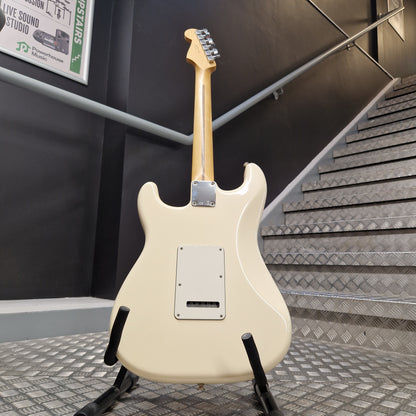 Pre-Owned Fender Standard Strocaster 2014 Electric - Arctic White Cream-colored electric guitar body on a stand, photographed in a real indoor environment with a staircase and dark wall in the background.