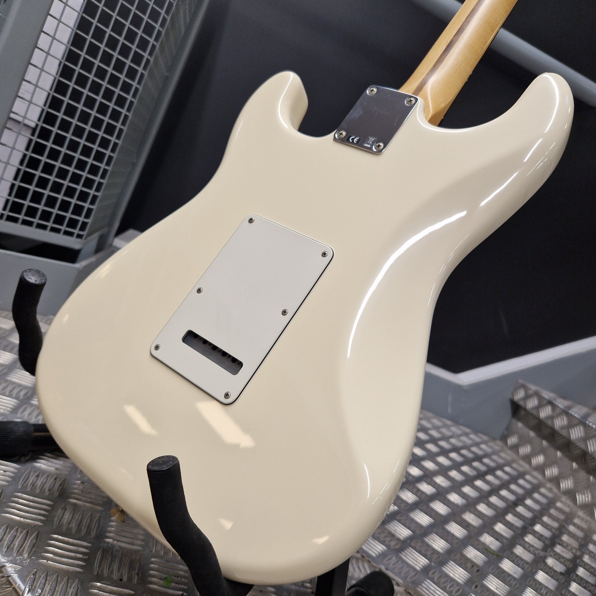 Pre-Owned Fender Standard Strocaster 2014 Electric - Arctic White Close-up of the rear body of an electric guitar in a glossy cream finish, mounted on a black display stand. The guitar features a silver pickguard and a light wood neck visible at the top, set against a dark workshop background with a metal grate.