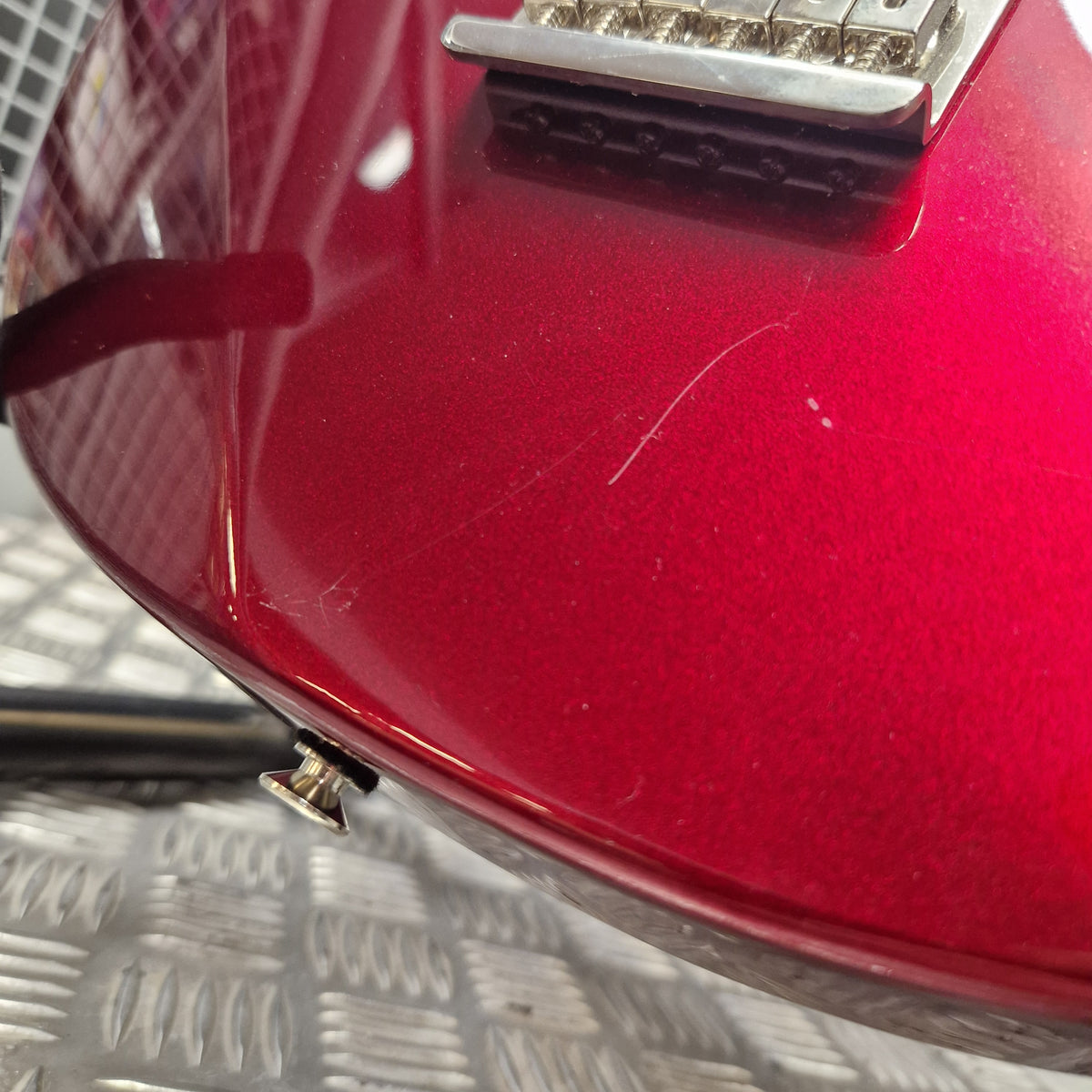 Pre-Owned Yamaha Pacifica 311H Electric - Red Metallic Close-up of a glossy red electric guitar body showing a chrome bridge, a small silver control knob, and some surface scratches on the finish, set against a textured gray metal surface.