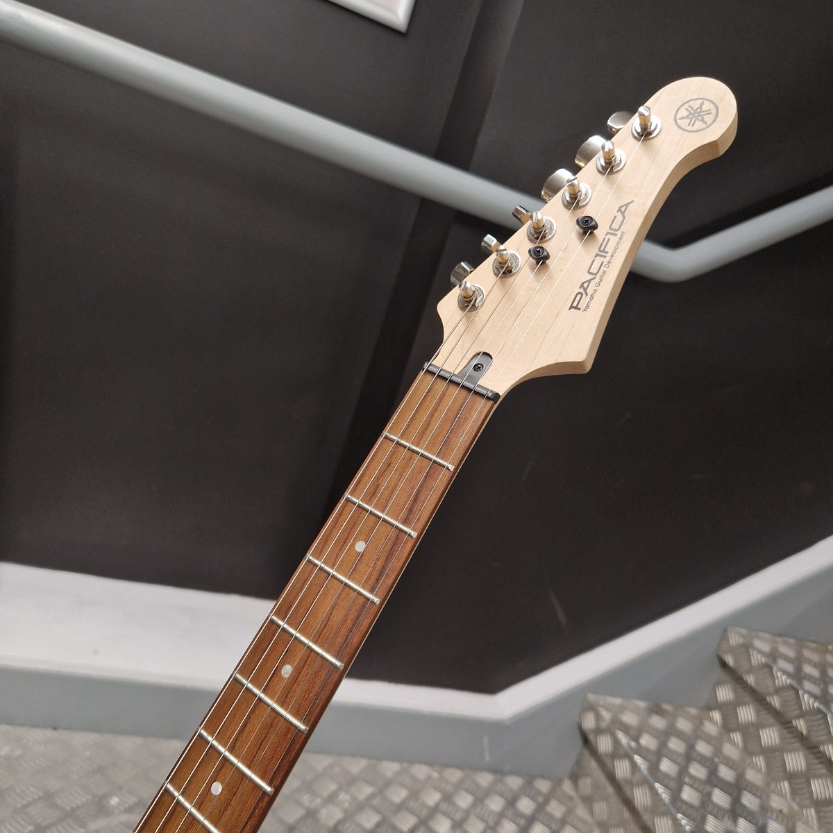 Pre-Owned Yamaha Pacifica 311H Electric - Red Metallic Close-up of a Yamaha electric guitar neck and headstock, showing the light natural wood headstock, silver tuning pegs, and dark brown fretboard against a dark gray staircase background.