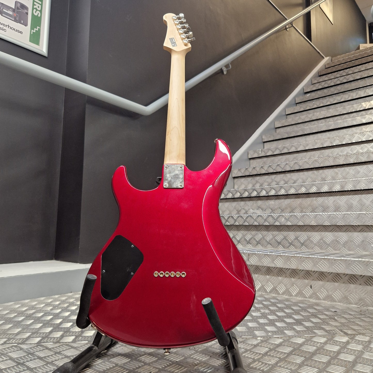 Pre-Owned Yamaha Pacifica 311H Electric - Red Metallic A red electric guitar with a natural wood neck and black pickups, displayed on a stand in front of a metal staircase.