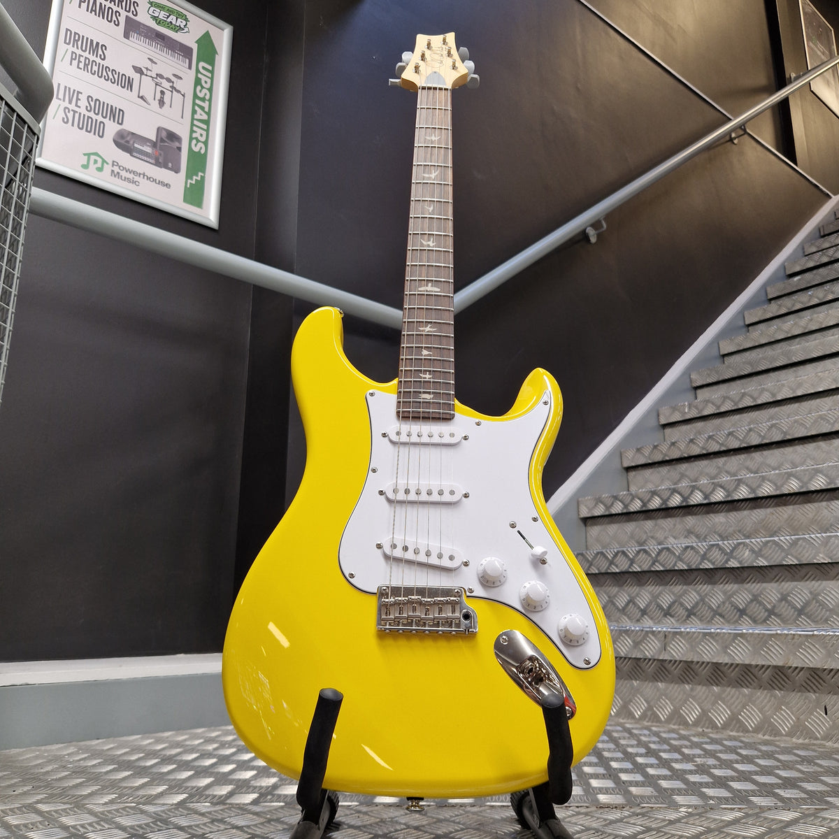 Pre-Owned PRS SE Silver Sky John Mayer Signature Electric - Dandy Lion A bright yellow electric guitar with a white pickguard and chrome hardware, displayed on a stand in front of a dark wall and staircase.