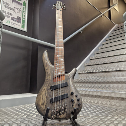 Pre-Owned Ibanez SRFF806 Multi-Scale 6-String Bass - Black Stained A double-cutaway electric bass guitar with a dark wood grain finish stands on a stand in front of a staircase.