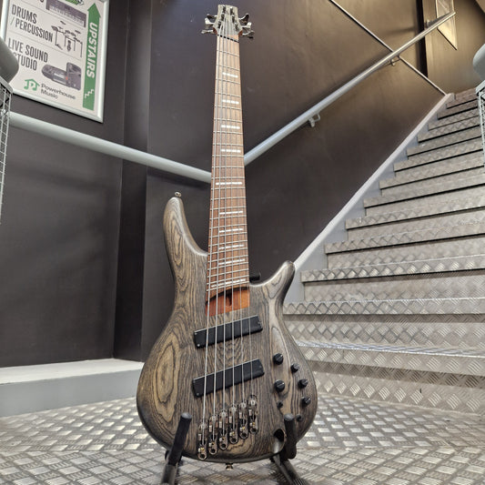 Pre-Owned Ibanez SRFF806 Multi-Scale 6-String Bass - Black Stained A double-cutaway electric bass guitar with a dark wood grain finish stands on a stand in front of a staircase.