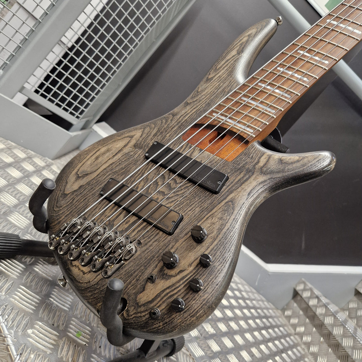 Pre-Owned Ibanez SRFF806 Multi-Scale 6-String Bass - Black Stained A close-up of a double-cutaway electric guitar with a dark brown wood-grain finish, resting on a metal stand in what appears to be a workshop or showroom.
