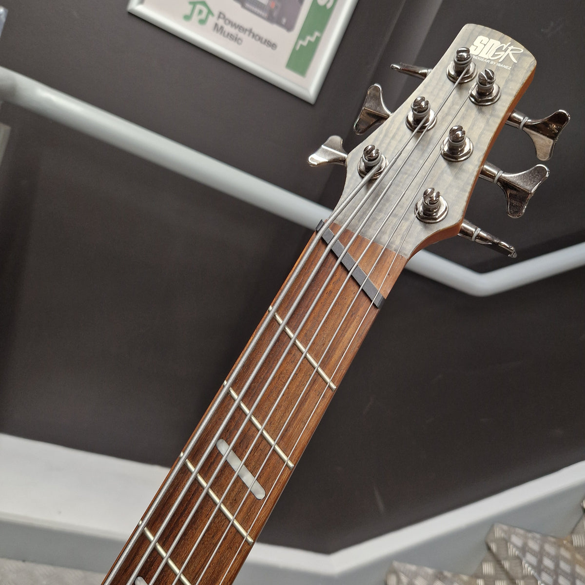 Pre-Owned Ibanez SRFF806 Multi-Scale 6-String Bass - Black Stained Close-up of a guitar headstock and fretboard, showing six tuning pegs, six strings, a maple neck, and a dark gray wall background.