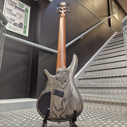 Pre-Owned Ibanez SRFF806 Multi-Scale 6-String Bass - Black Stained A custom electric bass guitar with a weathered, distressed wood grain finish, standing on a black stand in front of an indoor staircase.