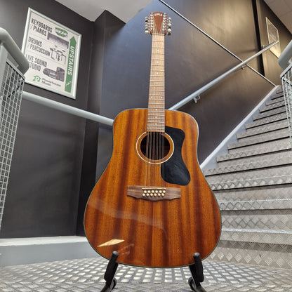 Pre-Owned Guild D-125-12NAT Dreadnought 12-String Acoustic - Mahogany A wooden acoustic guitar with a glossy brown finish and black pickguard, displayed on a stand in front of a staircase.