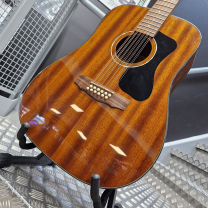 Pre-Owned Guild D-125-12NAT Dreadnought 12-String Acoustic - Mahogany A close-up of an acoustic guitar body with a glossy reddish-brown wood finish, black pickguard, and six-string tuning system, displayed on a stand in a workshop setting.
