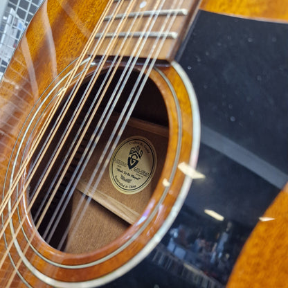 Pre-Owned Guild D-125-12NAT Dreadnought 12-String Acoustic - Mahogany Close-up of an acoustic guitar's sound hole and neck, showing a round gold-toned logo sticker in the center of the hole. The guitar has a glossy brown wood finish with a black pickguard and silver strings.