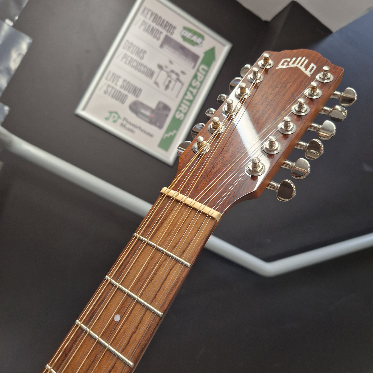 Pre-Owned Guild D-125-12NAT Dreadnought 12-String Acoustic - Mahogany Close-up of a wooden guitar headstock with 12 silver tuning pegs and a brown fretboard, photographed indoors against a dark background with a framed sign blurred behind it.