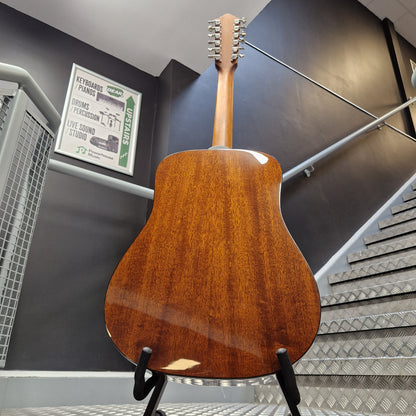 Pre-Owned Guild D-125-12NAT Dreadnought 12-String Acoustic - Mahogany A wooden acoustic guitar shown from the back, resting on a black stand in an indoor setting with a staircase and dark gray walls in the background.