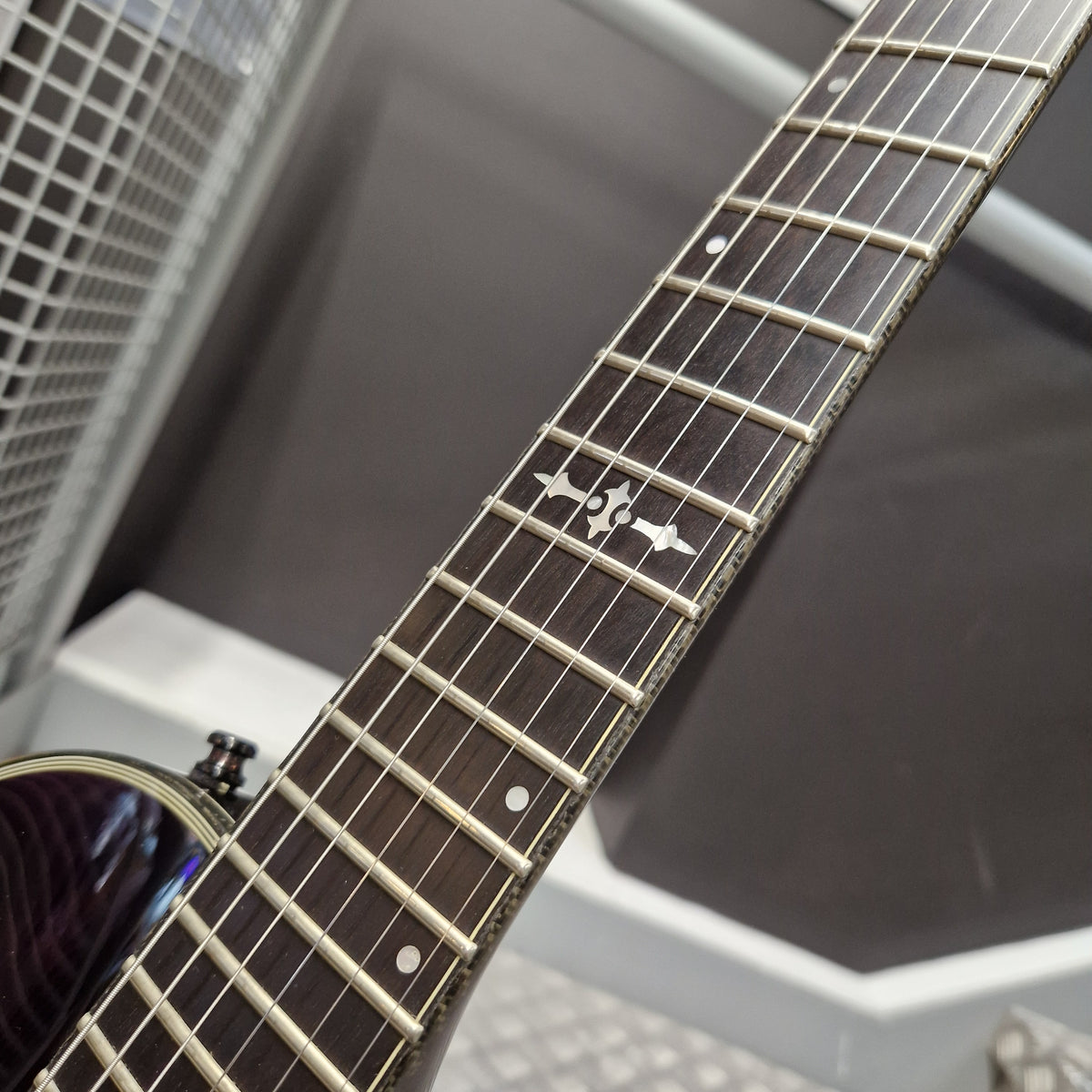 Pre-Owned Schecter Hellraiser Hybrid Solo-II Electric - Transparent Purple Burst Close-up of an electric guitar neck with a dark wood fretboard, silver metal frets, and a silver inlay featuring a stylized bird logo, set against a gray background.