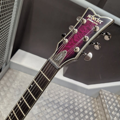 Pre-Owned Schecter Hellraiser Hybrid Solo-II Electric - Transparent Purple Burst Close-up of a Schecter electric guitar headstock and upper neck, showing a dark purple-to-pink gradient finish, chrome tuning pegs, and the Schecter logo on a gray metal floor.