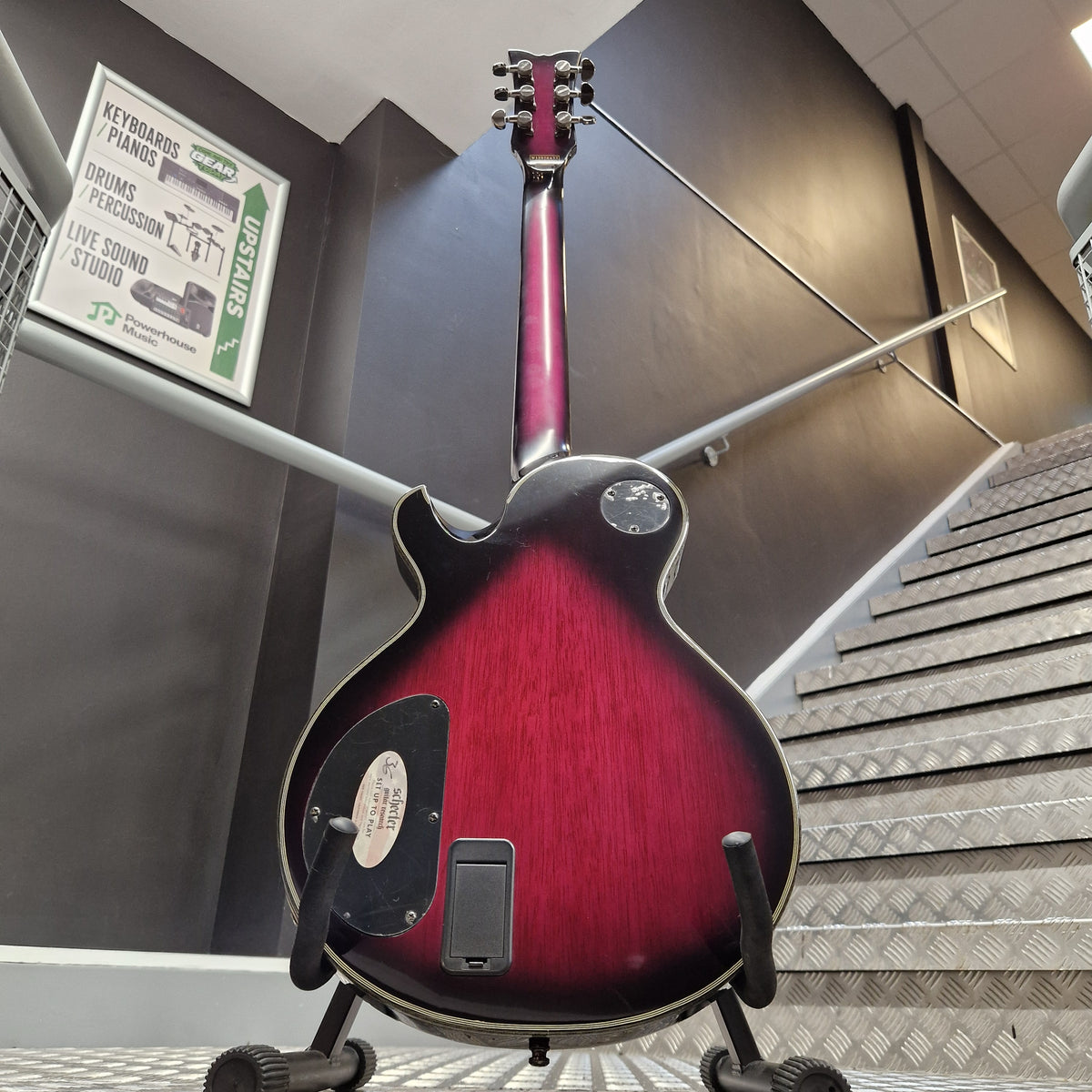 Pre-Owned Schecter Hellraiser Hybrid Solo-II Electric - Transparent Purple Burst A double-cutaway electric guitar with a dark red to black sunburst finish, displayed on a stand in an indoor setting with a staircase and grey walls in the background.