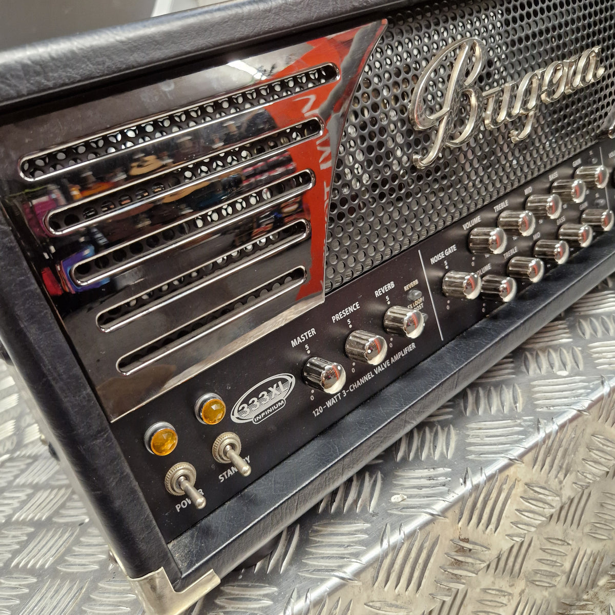 Pre-Owned Bugera 333XL Infinium 120w Electric Amplifer Head Close-up of a Buggio guitar amplifier head with chrome and black controls, silver grille, and a red-and-black graphic panel, resting on a diamond plate metal surface.