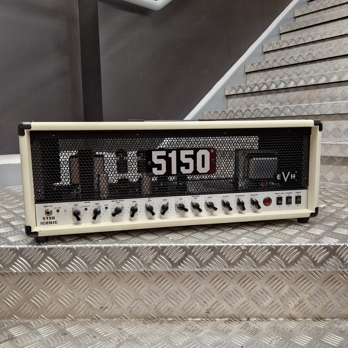 Pre-Owned EVH 5150 Iconic 80W Electric Amplifier Head A vintage-style electric guitar amplifier head sits on a metal diamond-plate staircase step in front of a dark wall. The unit has a cream-colored front case, a black mesh grille, large white '5150' text, and multiple black control knobs along the bottom.
