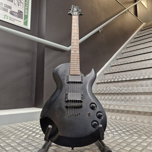 Pre-Owned Cort EVL-Z4 Electric - Satin Black A black electric guitar with a dark wood fretboard and black hardware, displayed on a stand in front of a staircase.