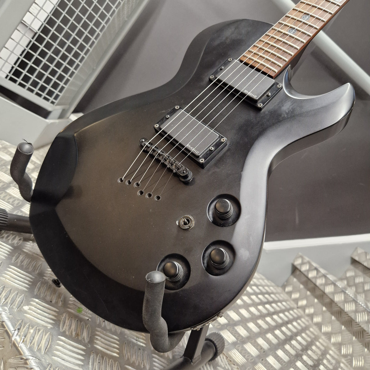 Pre-Owned Cort EVL-Z4 Electric - Satin Black A close-up of a black electric guitar body with dual humbucker pickups, three control knobs, and a dark fretboard, resting on a metal stand.