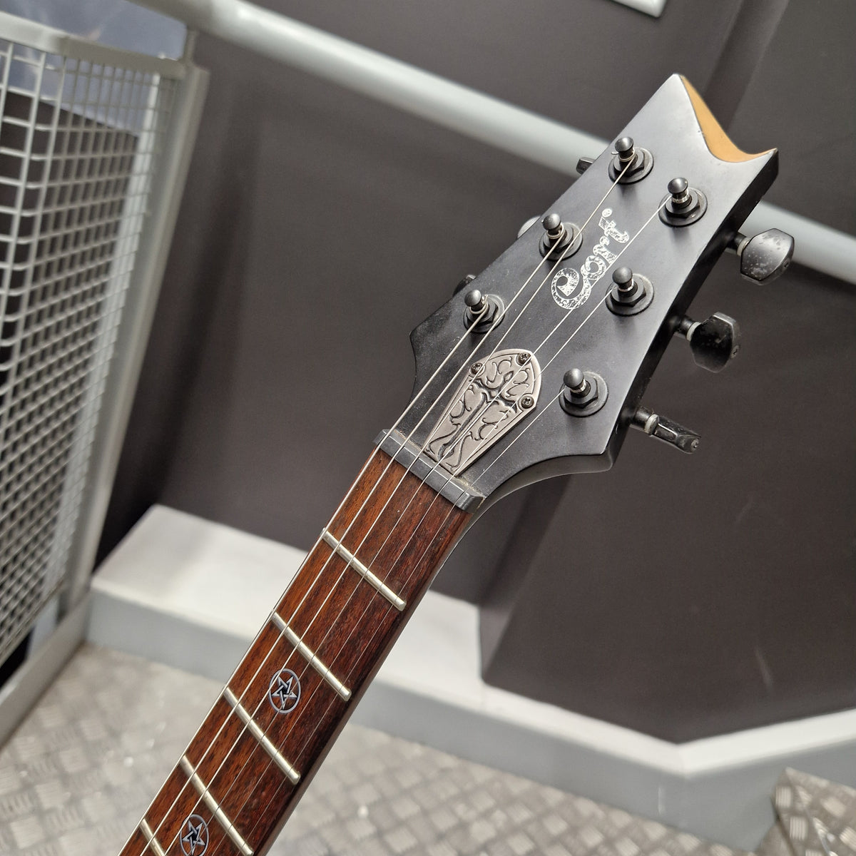 Pre-Owned Cort EVL-Z4 Electric - Satin Black Close-up of a guitar headstock and fretboard, showing a dark wood neck, black headstock with silver hardware, and a gray indoor background.