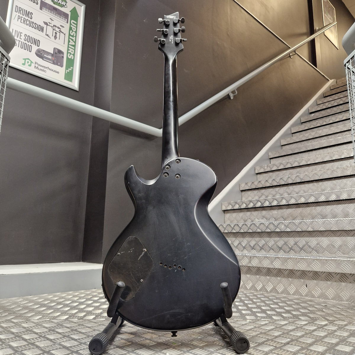 Pre-Owned Cort EVL-Z4 Electric - Satin Black A black electric guitar body with a distressed finish, displayed on a stand in front of a staircase.