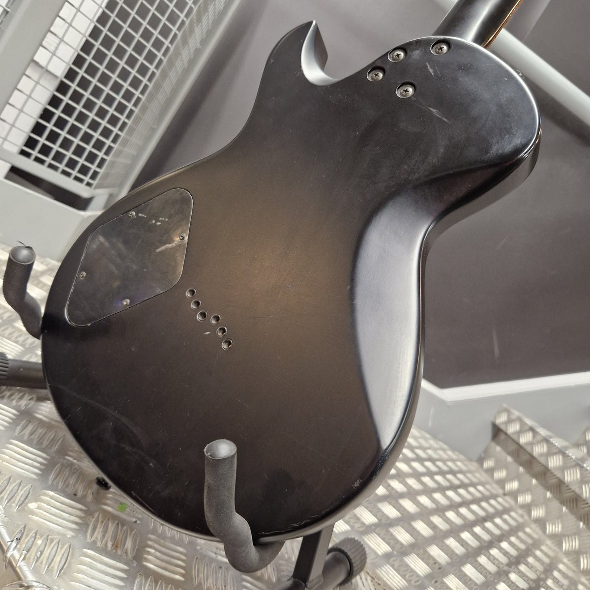 Pre-Owned Cort EVL-Z4 Electric - Satin Black A used black electric guitar body, photographed from the back, resting on a metal stand on a diamond plate floor.